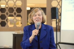 Woman holding a microphone and speaking during an indoor community presentation.