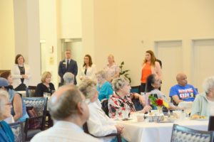 Residents seated at tables listening to staff during a community gathering.