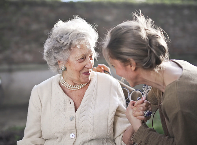 Older woman seated outdoors holding hands with a younger woman while they talk closely.