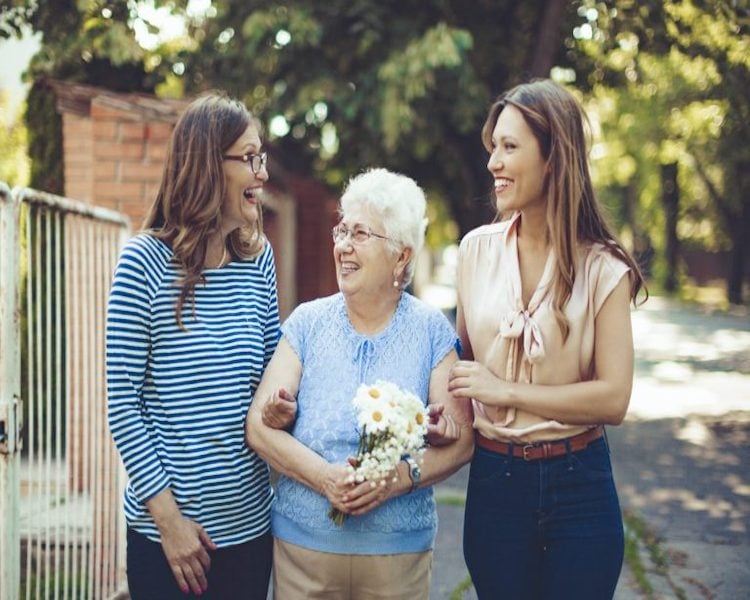 Older woman walking outdoors with two younger women, smiling and holding flowers.