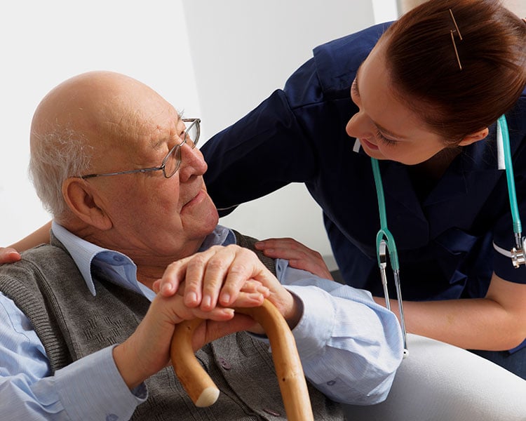 Caregiver assisting an older man seated with a walker during a personal care interaction.